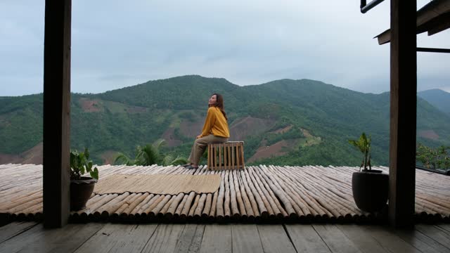 A young woman walking into balcony and sitting on wooden chair, looking at a beautiful mountains view