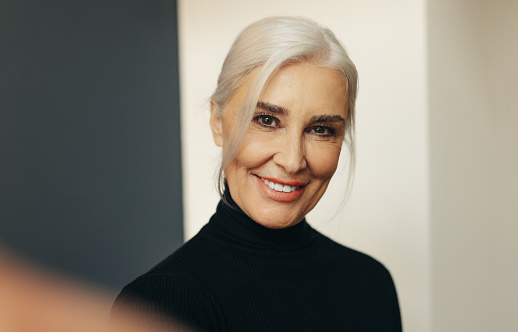 Senior professional woman with silver hair flashing a warm and genuine smile as she takes a selfie, expressing confidence and pride in her years of business experience.