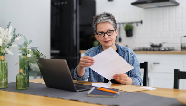 istock Senior woman at a table with a laptop in a modern interior holds reports by mail, documents in her hands. 1499707483