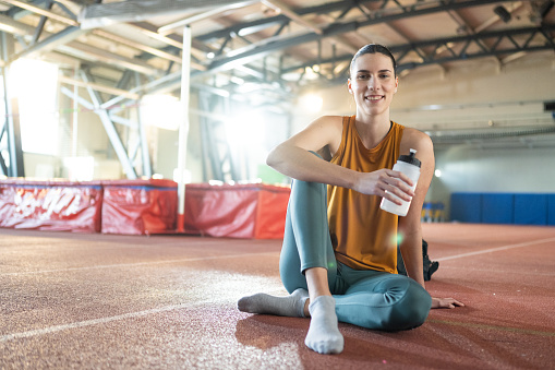 Portrait of smiling athlete young woman holding water bottle while sitting in sports hall.