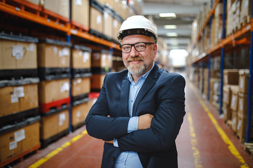 Portrait of a happy attractive business manager man wears white protective helmet and black suit standing in the aisle and controls huge shelves around boxes, industrial equipment and materials for work in a new stock factory distribution warehouse.