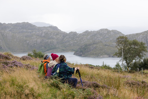 A medium wide angle view of three senior women, taking in the fantastic view whilst out hiking in the Scottish highlands. They have stopped to have a little sit down and take a moment to remember this memory. They are dressed appropriately for the cooler temperature and I using hiking poles on their journey.