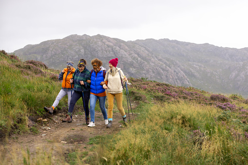 A wide angle view a group of mature women all getting back to nature and enjoying a trip to the Scottish highlands where they are hiking together and admiring the picturesque views along the way. The weather is perfect for a hike and they are linking arms as they reach a peak along the trail.