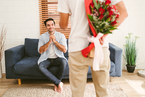 Young gay couple looking at the boyfriend, holding a bouquet of roses behind his back for the surprise at home. LGBT couple surprises boyfriend with a rose bouquet. LGBT couple living together concept