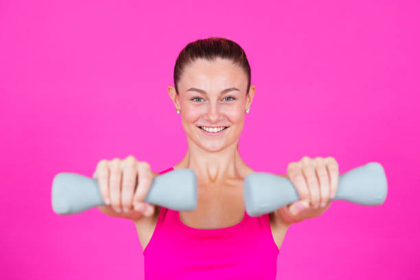 Young pretty sportswoman holding weights and looking at camera isolated on pink background.
