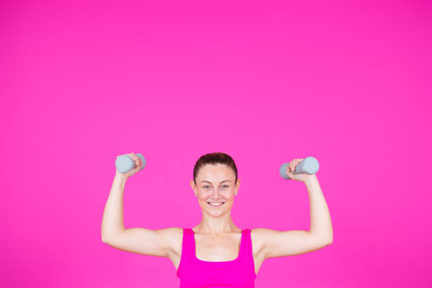 Smiling strong sportswoman lifting weights isolated on pink background with copy space. young woman looking at camera.