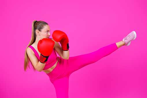 Fighter woman wearing boxing gloves and giving a kick isolated on vibrant pink background. Feminism and woman power concept.