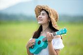 Lifestyle asian women white dress holding a guitar on a cloudy sunrise sky in the meadow flower, relax and happy day on summertime, outdoors background field copy space for inscription.