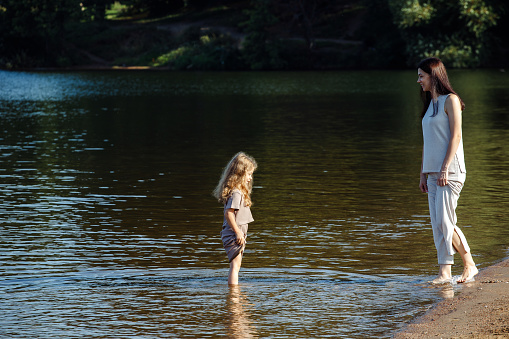 A five-year-old cheerful girl and her mother are walking by the lake, barefoot in the water, having fun on a summer day. Happy time.