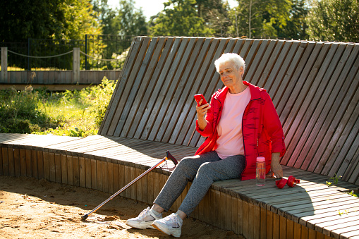 An elderly woman with short gray hair sits on a wooden bench in the park and watches the news on a smartphone, lifestyle concept.