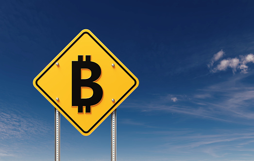 Bitcoin symbol written yellow off road traffic sign before clear blue sky. Horizontal composition with copy space.