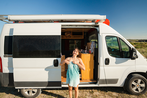Young Caucasian woman in a towel standing near the camper van after swimming in the sea