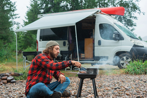 Young Caucasian man in red checked shirt barbecuing near the river and camper van  in Norway