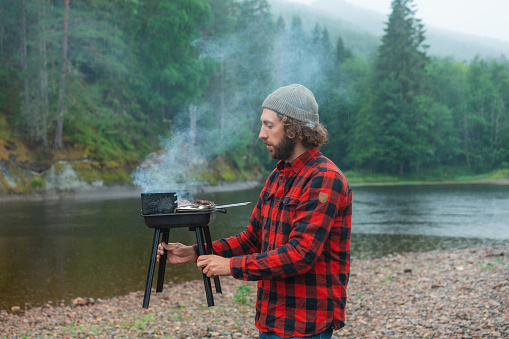 Young Caucasian man in red checked shirt barbecuing near the river in Norway