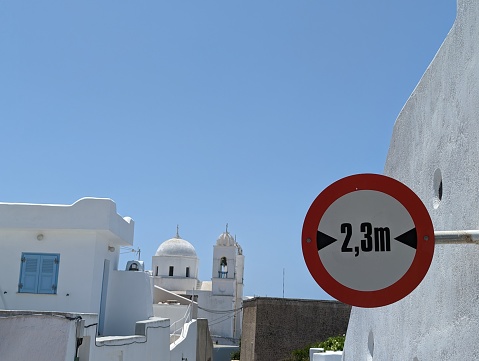 White washed buildings and church in a Greek village with a street sign in the foreground warning of a narrow street