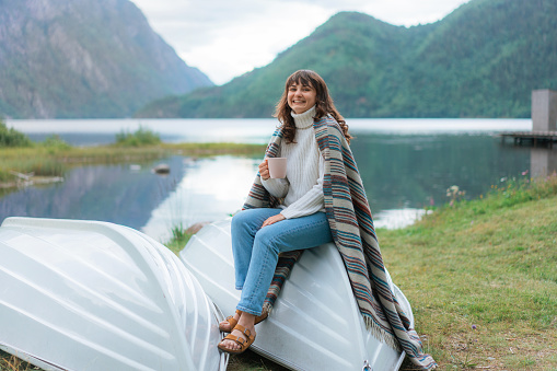 Young Caucasian woman in white sweater sitting on the boat near the lake and drinking tea