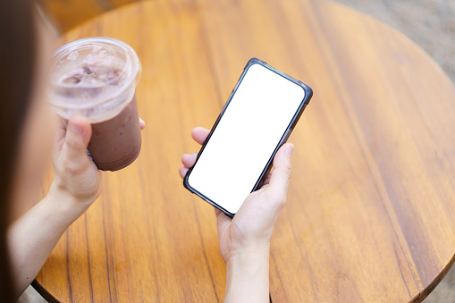 Isolated with clipping path of empty white screen of smartphone. Beautiful good looking Asian woman drinking an iced chocolate drinks in the glass, woman relaxing in the cafe or coffee shop and using smartphone.