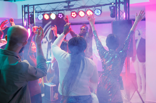 Energetic crowd dancing and enjoying discotheque on dancefloor with colorful spotlights. People jumping, having fun and moving to music beats with raised hands in nightclub