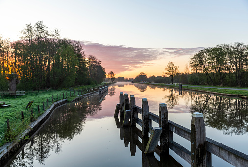 Calm canal at dusk. Romantic sky reflecting on water surface. Silhouettes of trees, yarrow and grass. Taken with long exposure at Ems-Jade-Kanal in Sande, Friesland, Lower Saxony, Germany, Europe.