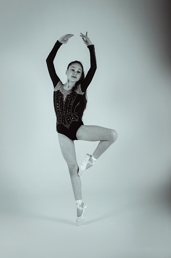A girl gymnast in a black gymnastic leotard stay in a graceful pose in pointe shoes. White nd black. Front view