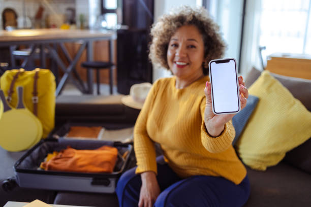 Happy mature woman packing for a journey and showing smart phone with blank screen stock photo