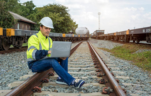 Male railway engineer in reflective green work clothes and white helmet uses laptop computer while sitting on railway tracks.
