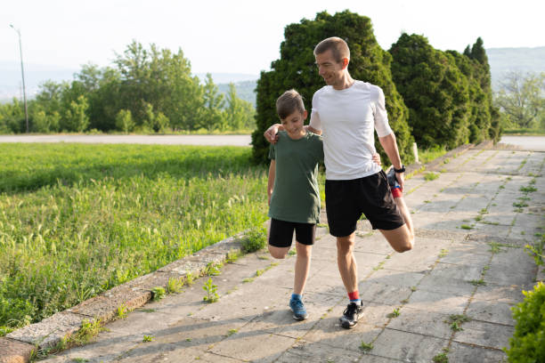 A man in a blue shirt reaches out to his friend who is kneeling to tie his shoelaces on a park pathway, bordered by trees and basking in the sunlight.
