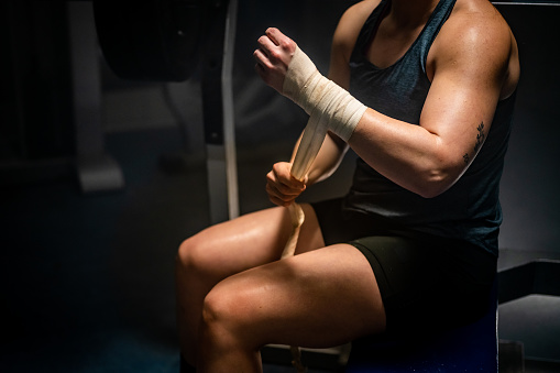 Athlete woman wrapping hands with arms bandage while sitting on bench in gym.