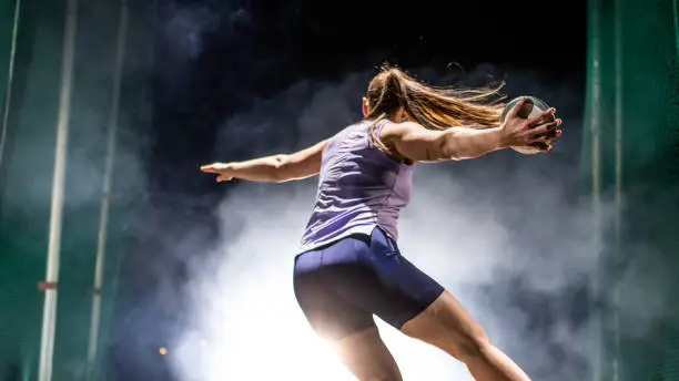 Athlete woman throwing discus on track during practice at night.