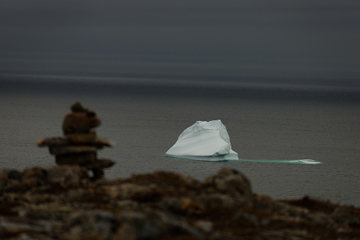 Single medium sized iceberg floating off of the coast of Newfoundland and Labrador. Rocky cliff and Inuksuk in foreground.