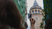 tourist woman examining the photo of galata tower, comparing galata tower in reality and photo, beautiful woman taking street photo among colorful houses, young tourist woman doing street photography with pink camera, retro street photography
