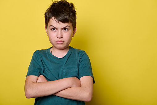 Isolated close-up emotional portrait on yellow background of a teenager boy, elementary school student with arms folded, looking at camera with sad, unhappy and sorrowful gaze. Back to school.