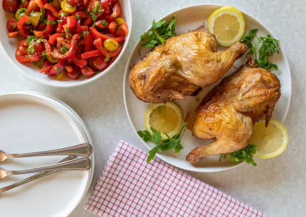 Delicious homemade oven roasted or baked half chicken. Served with a tasty and healthy tomato, bell pepper salad on white table background with plates and cutlery. Top view