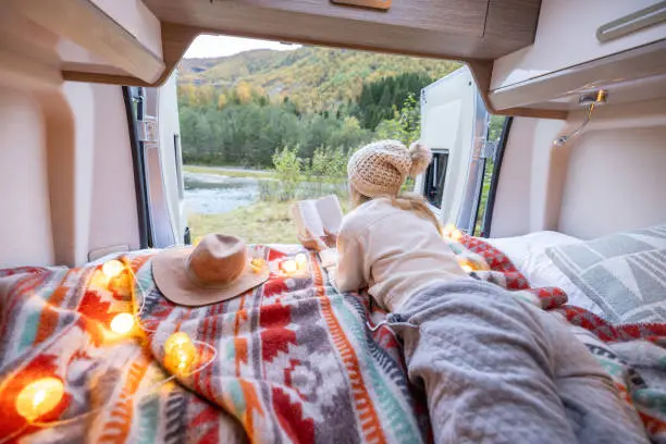 Photo of Woman relaxing at the back of her camper van reading a book, van life concept.