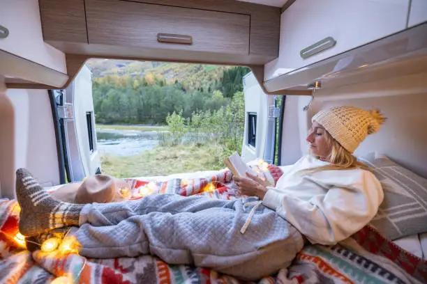 Photo of Woman relaxing at the back of her camper van reading a book, van life concept.