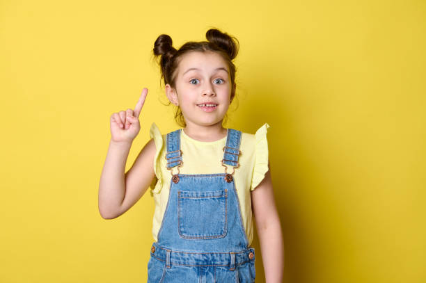 retrato de estudio asombrado niña apunta con el dedo índice hacia arriba, mostrando un espacio publicitario de copia sobre fondo amarillo aislado - brain gym fotografías e imágenes de stock
