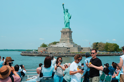 New York City, United States - May 22, 2023: A tour boat full of tourists browses in front of the Statue of Liberty, in Liberty Island, New York, United States, on a sunny spring day
