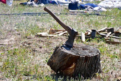 A close up on an old medieval axe used for chopping wood seen next to some planks, boards, and logs with the edge of the tool being inside of a big old rusty log used as a chopping block