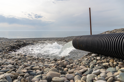 Close-up of a large plastic water pipe with a large stream of water gushing onto a pebble beach against the background of the sea