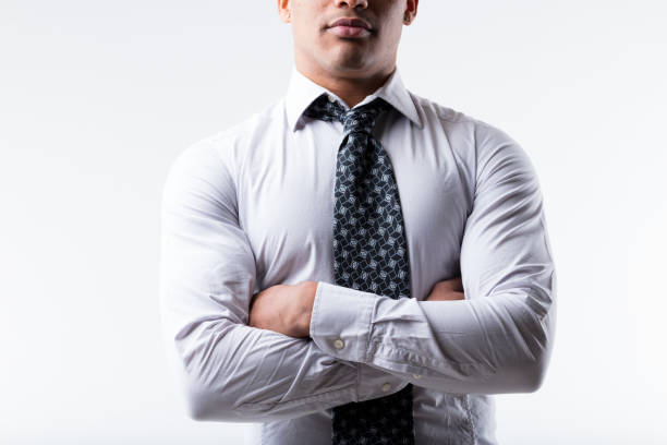 Businessman of black ethnicity, partially visible face, stands arms crossed, exuding strength and steadfastness. In white shirt, dark patterned tie. Muscular, white background