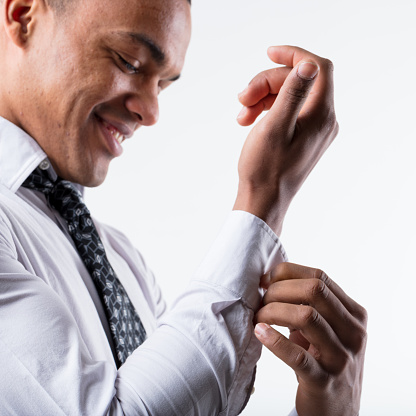 Sharp detail of a young black man's hands buttoning up an elegant white shirt. A businessman dedicated to his job or a groom preparing for his bride? His slightly blurred face smiles widely