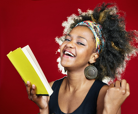 Young woman with multi-colored hairband and natural black hair looks joyful as she holds a book she's reading.