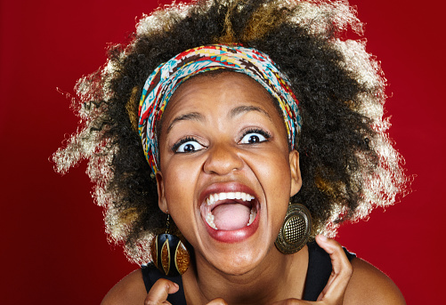 Young woman with multi-colored hairband and natural black hair shouts as she reacts in some combination of excitement and frustration.