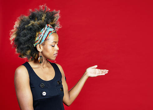 Young woman with multi-colored hairband and natural black hair uses her hand to draw attention to copy space on red background.