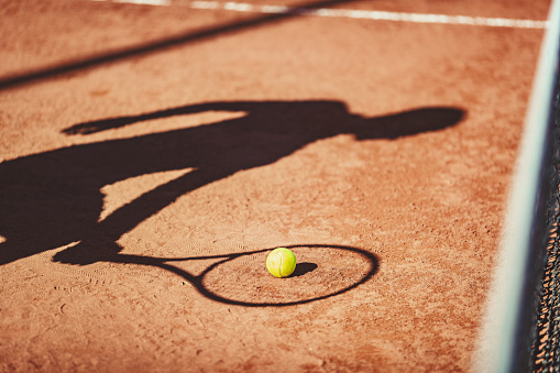 Shadow of a tennis ball and tennis player in action on a clay court .