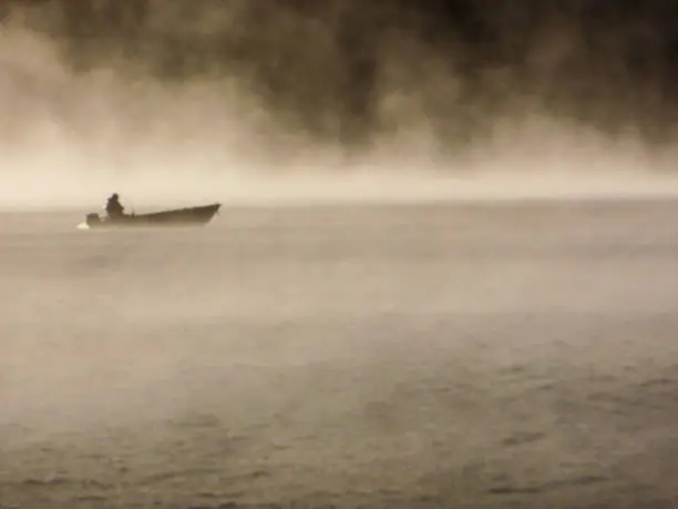 A lone fisherman in a boat drifts amongst mysterious rising mist on Whiteswan Lake