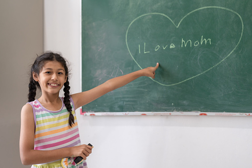 Happy cute girl using chalk drawing heart and write love mom on chalkboard wall in classroom at elementary school. Children education and learning concept.