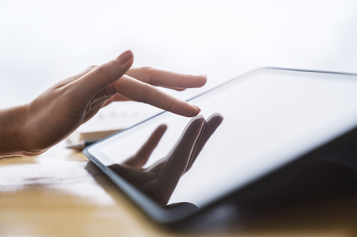 Close detail of a woman's finger making contact with the screen of a sleek digital tablet resting on a desk, set against the soft-focus backdrop of a light-filled office