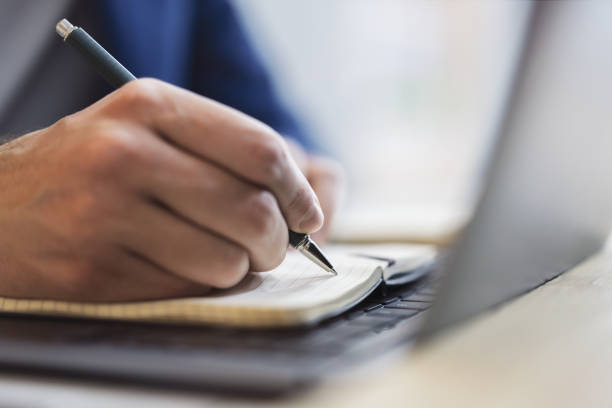 istock Detailed view of a man's hand jotting down notes in a notepad on a modern laptop, with a blurred background 1499801660