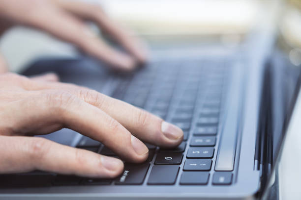 クローズアップは、柔らかくぼかされたオフィスの背景に設定された、現代のラップトップのキーボードで入力する男性の手を示しています - mail keyboard button ストックフォトと画像